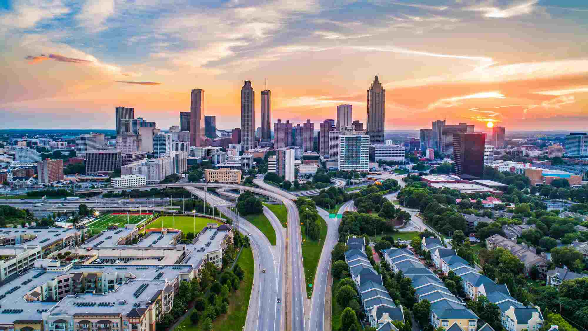 Aerial view of downtown Atlanta, Georgia at sunset, showing high-rise buildings and major highway intersections surrounded by green spaces and residential areas.