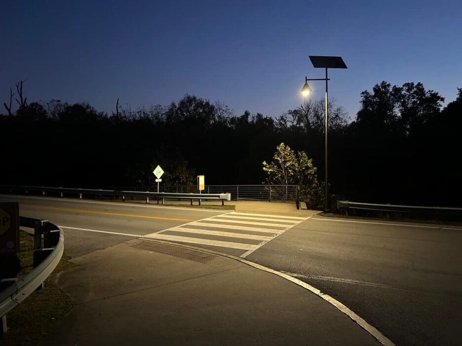 Solar-powered streetlight illuminating a quiet crosswalk at dusk near a wooded roadway.