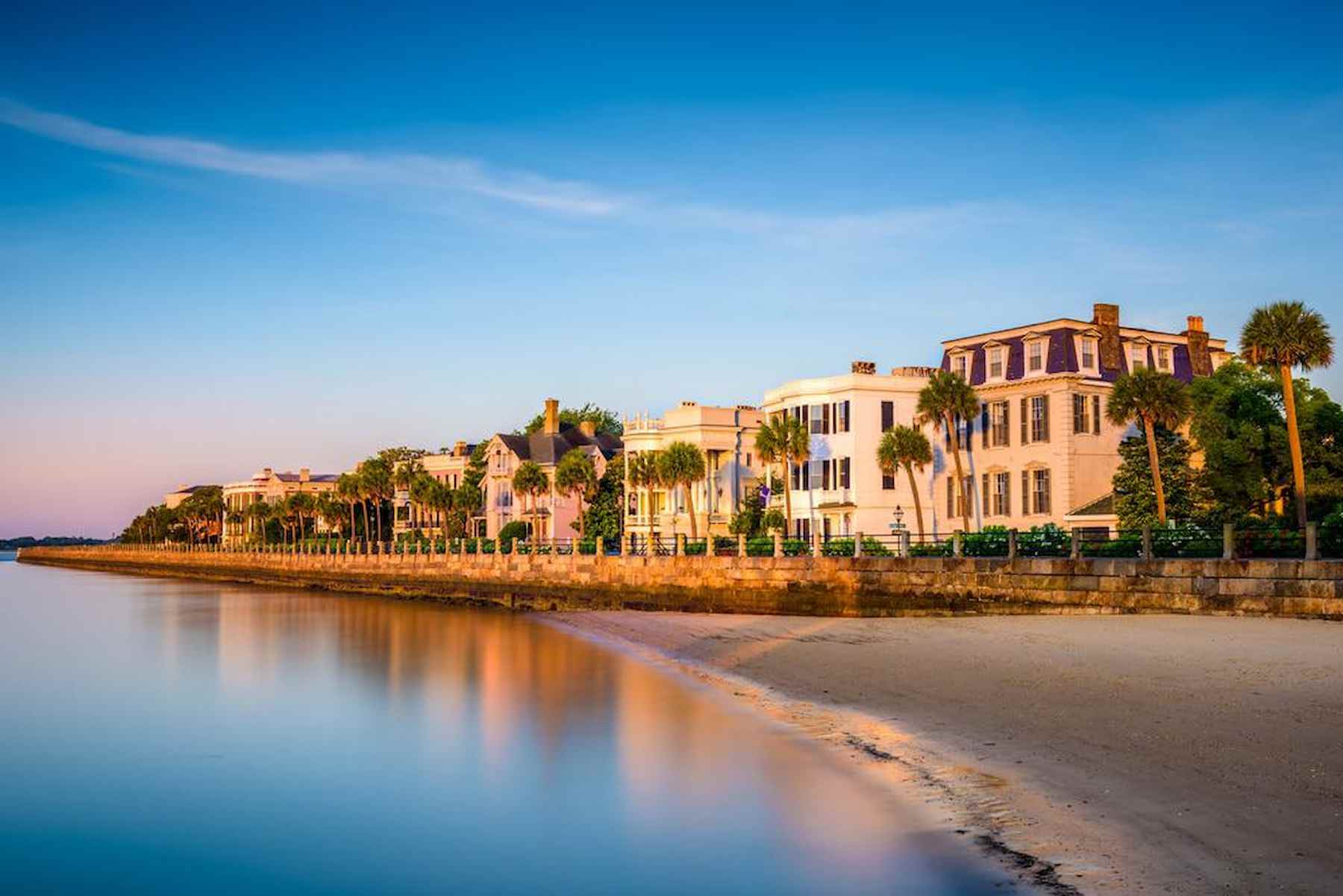 Historic waterfront homes along The Battery in Charleston, South Carolina, at sunrise, with palm trees lining the promenade and calm ocean waters in the foreground.