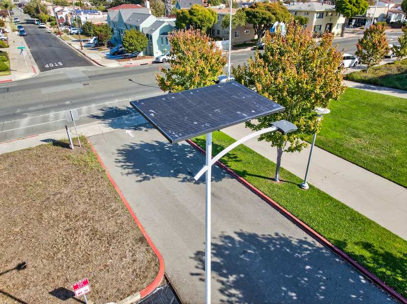 Solar-powered streetlight installed on a residential roadway with adjacent sidewalks and green space &mdash; no trenching, no grid connection required.
