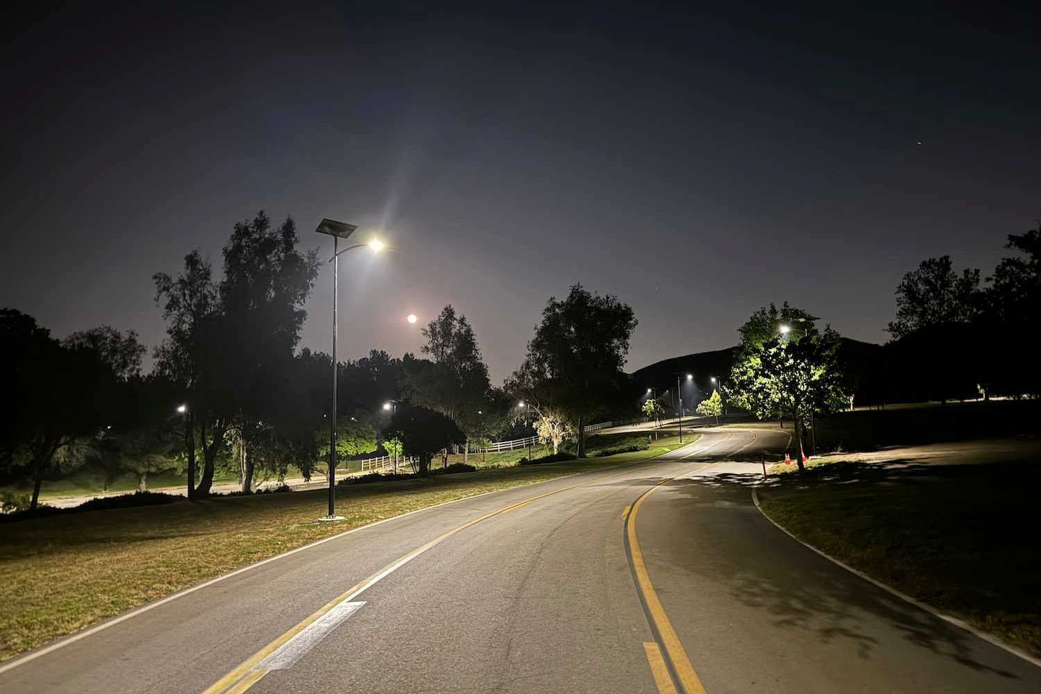 Curved park roadway illuminated by solar streetlights at night, with trees and a visible moon enhancing the serene landscape.