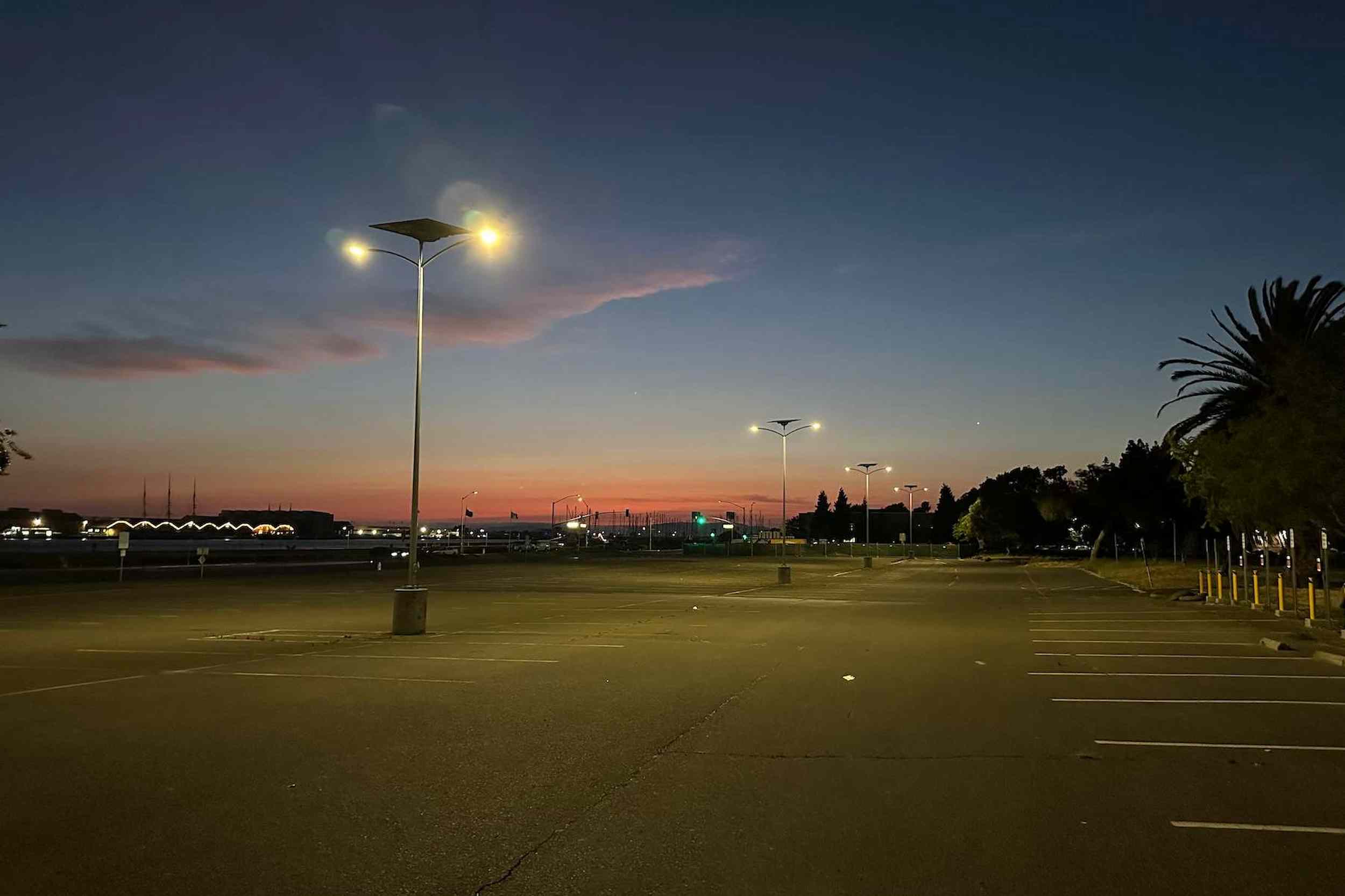 Large empty parking lot illuminated by solar-powered streetlights at dusk, with palm trees and cityscape in the background.