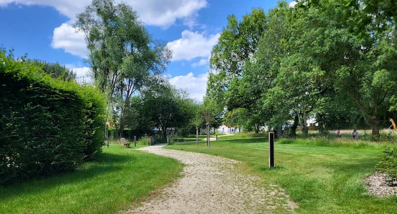 NOWATT solar bollard installed along a sunlit park path, providing clean, off-grid lighting in a greenway with trees and open sky.