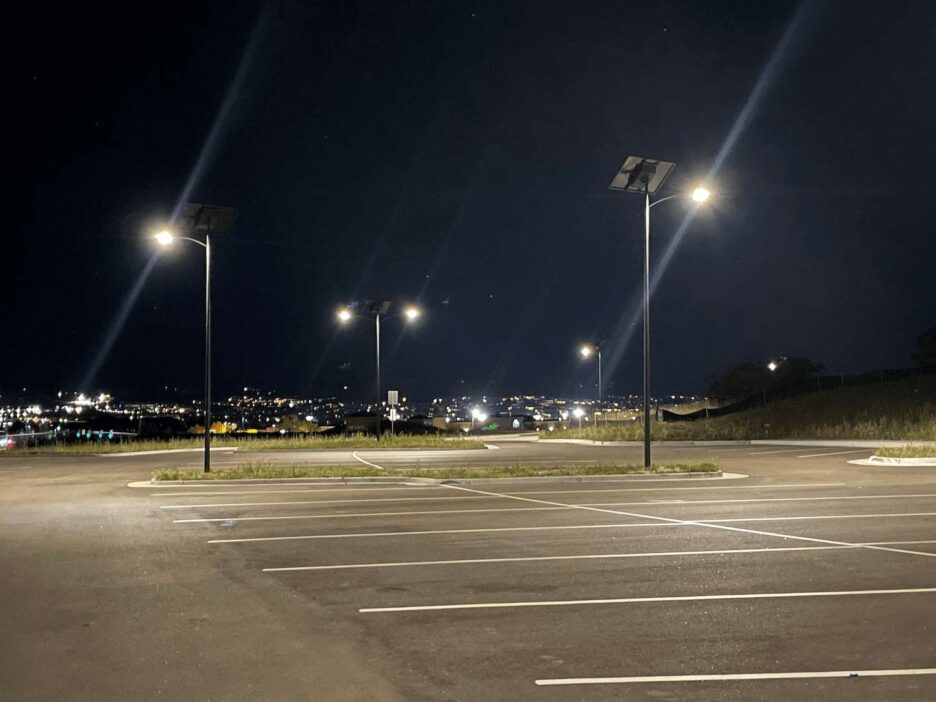 Solar-powered streetlights illuminating an empty parking lot at night with city lights visible in the distance.