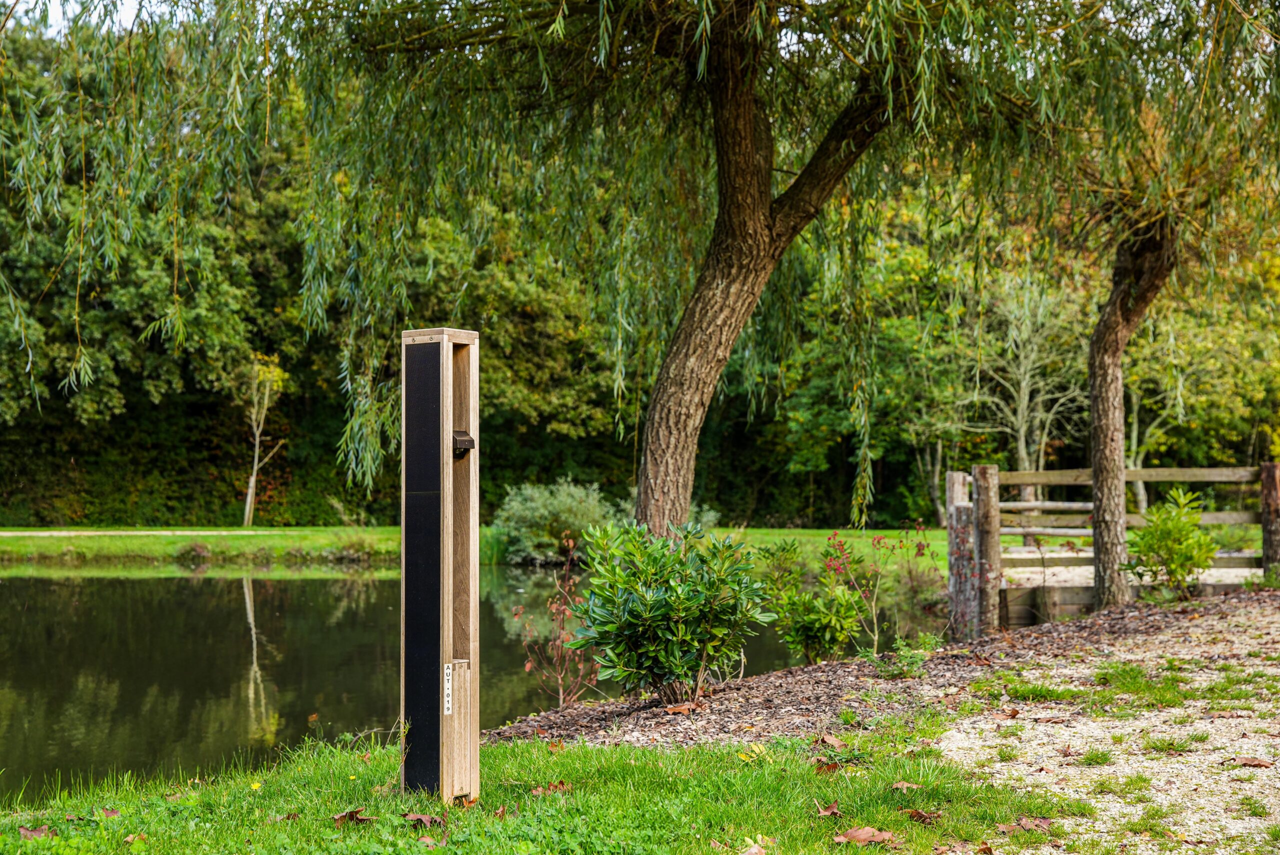 Sustainable wooden NOWATT solar bollard installed near a pond and walking trail, offering off-grid architectural lighting that blends naturally into landscaped environments.
